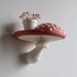 3D printed wall shelf shaped like a red and white amanita mushroom with a flat spotted cap and sculpted gills, used as a small floating ledge holding a vase of flowers.