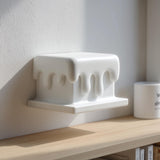 3D printed wall shelf shaped like a rectangular block with smooth dripping edges made of matte PLA plastic, mounted above a wooden shelf in soft natural daylight.