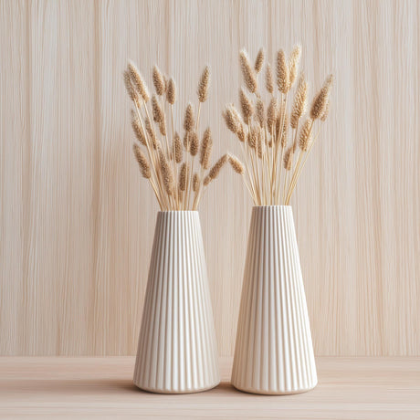 Two cream-colored origami-style vases with a pleated texture, each holding dried grasses, displayed against a light wooden background.