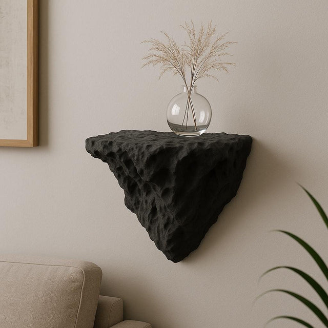 A black 3D printed wall-mounted shelf with a rugged, rock-like texture and triangular shape, displayed in a minimalist interior. The shelf holds a round glass vase with dried ornamental grass, and is mounted above a beige couch against a soft-toned wall.