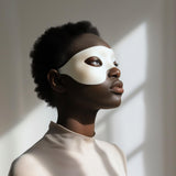 Adult person wearing a minimalist white 3D printed eye mask, standing in soft natural light against a neutral interior background.