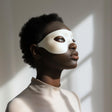 Adult person wearing a minimalist white 3D printed eye mask, standing in soft natural light against a neutral interior background.