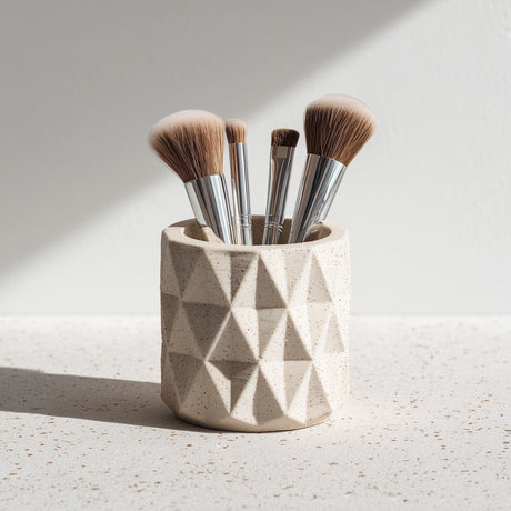 A beige 3D printed geometric cup holding makeup brushes, shown on a speckled surface with natural light. The cup features a faceted, textured design and matte finish.