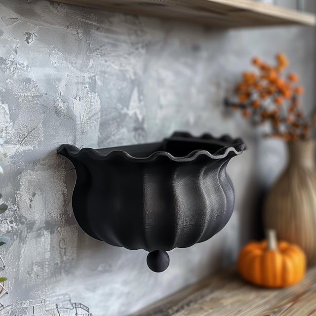 3D printed wall shelf shaped like a black witch cauldron with wavy rim, matte PLA texture, mounted on a textured grey wall in a cozy autumn interior.