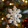 3D printed white geometric snowflake Christmas tree ornament hanging from a branch among warm fairy lights and festive baubles.
