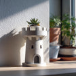 3D printed planter shaped like a small castle tower or rook, holding a succulent on a windowsill, with soft natural light and other plants in the background.