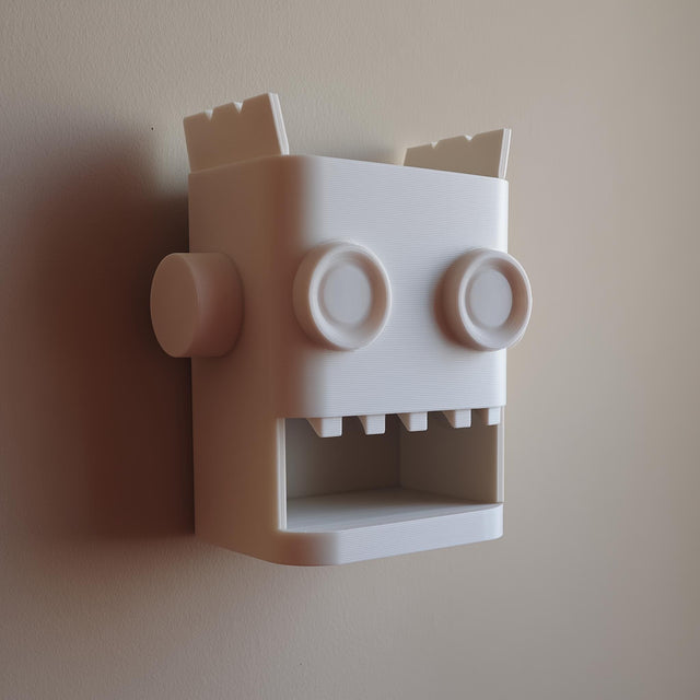 White 3D printed robot head wall organizer with an open mouth storage niche, made of matte PLA and mounted on a plain wall in soft natural light.