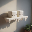 3D printed wall shelf with a dripping edge and two marshmallow-like cube characters with faces, made of matte PLA plastic and mounted on an interior wall in natural light.