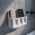 3D printed white wall-mounted organizer shelf with two upper compartments for phone and remote and three small front pockets, installed above a bathroom sink on a textured grey wall.