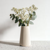 A tall, cream-colored 3D printed origami vase with vertical ribbed lines, holding white dried flowers and eucalyptus stems, placed on a speckled countertop.