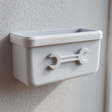 3D printed white wall-mounted organizer bin with an embossed wrench icon on the front, attached to a textured wall in a garage or utility room.