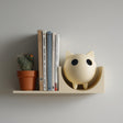 3D printed wall shelf with a round cat figure acting as a bookend beside books and a small cactus pot on a slim floating ledge against a light wall.