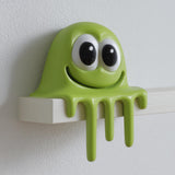 Cute 3D printed green slime monster figurine sitting on the edge of a flat shelf with big cartoon eyes and dripping goo front.
