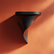3D printed matte PLA wall-mounted shelf with a round top and elegant twisted spiral bracket, in dark black, against a warm-toned wall.