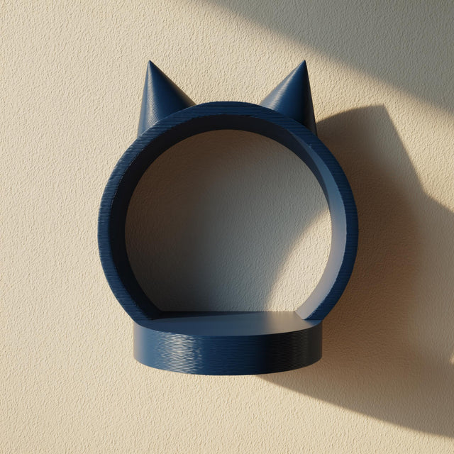 3D printed matte PLA wall shelf shaped like a round ring with small cat ears on top, in a solid dark blue color, mounted on a light wall in warm sunlight.