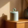 Cute minimalist 3D printed pet cremation urn with cat ears and curled tail lid handle, displayed on a wooden cabinet in warm light.