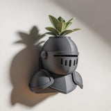 3D printed wall mounted knight helmet planter shelf with a small green plant, displayed on a light wall with soft natural shadows.