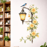 A vertical watercolor illustration of an antique dark metal street lamp on a white background. The lamp is lit with a warm yellow glow and is entwined with a vine of pale yellow roses and small white flowers. A bluebird with an orange breast sits on the curved arm of the lamp.