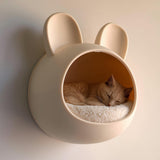 3D printed bunny-shaped wall mounted cat bed in matte beige PLA with a sleeping ginger cat inside, mounted on a warm neutral wall.