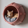 3D printed industrial barrel-shaped wall mounted cat bed in matte terracotta PLA with a ginger cat sleeping inside on a gray cushion.