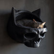3D printed dark fantasy skull wall mounted cat bed in matte black PLA with a sleeping tabby cat inside, gothic interior setting.
