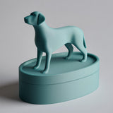 3D printed oval pet ashes urn with a standing dog figurine on the lid, clean minimalist keepsake container with visible print texture on a studio background.