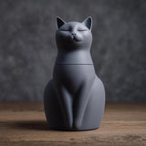 3D printed minimalist cat-shaped pet ashes urn with closed eyes and a fitted lid seam, visible print texture, on a wooden tabletop with a dark backdrop.