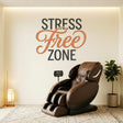 A wall decal that reads STRESS Free ZONE. The words STRESS and ZONE are printed in large, dark charcoal grey block letters. The word Free is positioned between them in an elegant, flowing, terracotta-colored cursive script. The text is applied directly to a smooth, light-colored wall, creating a clean, painted-on appearance. The surrounding settings include a plush brown massage chair, a modern office meeting room, and a serene yoga space, suggesting a peaceful, relaxing environment.