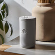 3D printed minimalist cylindrical pet ashes urn with a raised paw print emblem and fitted lid, displayed on a tabletop in soft natural light.
