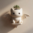 3D printed kawaii bat cat wall planter shelf in matte cream PLA, mounted on a warm neutral wall and holding a small succulent plant.