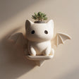 3D printed kawaii bat cat wall planter shelf in matte cream PLA, mounted on a warm neutral wall and holding a small succulent plant.