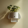 3D printed minimal dog-shaped wall planter shelf in matte white PLA mounted on a soft warm wall and holding a green houseplant.