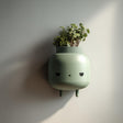 3D printed minimalist character wall planter in matte sage green PLA mounted on a neutral wall, holding small green plants.