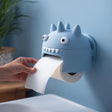 3D printed spiky kawaii monster wall mounted toilet paper holder with round eyes and teeth, hand pulling toilet paper from the mouth, bathroom sink background.
