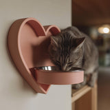 3D printed heart-shaped wall mounted pet bowl holder with a round bowl opening, shown with a cat eating from a metal bowl in a cozy home interior.