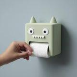 3D printed kawaii monster wall mounted toilet paper holder with small horns, round eyes, and teeth, with a hand pulling toilet paper from the mouth opening.