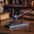3D printed raven skull magic wand with display stand in matte black PLA on a wooden desk surrounded by gothic fantasy decor.